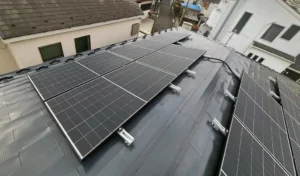 2_01 - 東京都の補助金制度を活用した太陽光・蓄電池・V2H設置サポート|ソルト公式 Row of dark solar panels mounted on a gray metal roof in an urban neighborhood viewed from above, showing wiring nearby