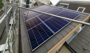 Blue solar panels mounted on a sloped residential roof with metal scaffolding nearby and neighboring houses in the background.